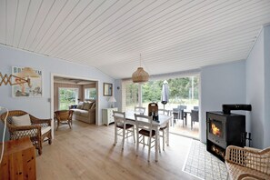 A spacious living area with a dining table, wicker furniture, a fireplace, and large windows. - Charmantes Sommerhaus in der Nähe eines fantastischen Sandstrandes (Åkirkeby)