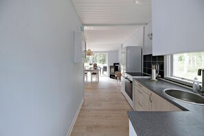 A modern kitchen with a large window, wooden flooring, and a dining area in the background. - Charmantes Sommerhaus in der Nähe eines fantastischen Sandstrandes (Åkirkeby)