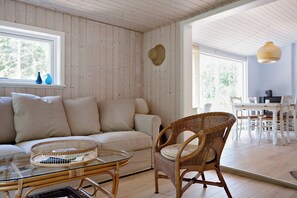 A room with a sofa, wicker chair, glass coffee table, and a dining area with a table and chairs. - Charmantes Sommerhaus in der Nähe eines fantastischen Sandstrandes (Åkirkeby)