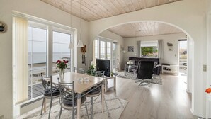 Ein großzügiger Wohnbereich mit Esstisch, Stühlen, einer Couch und einem Fernseher. Ein großes Fenster mit Vorhängen, eine Holzbalkendecke und Blick ins Freie.