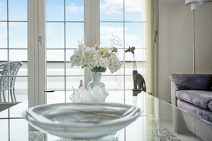 Un intérieur moderne avec une table en verre, des fleurs blanches et une vue dégagée sur l’extérieur grâce à de grandes fenêtres.