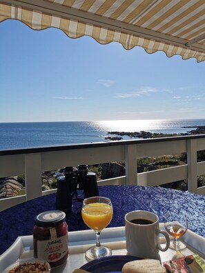 A table with a tray containing a jar of Marmalade, a glass of orange juice, a cup of coffee, and a glass of whiskey, set against a backdrop of a clear blue sky and a rocky coastline.