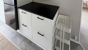 Et kompakt køkken med et indbygget køleskab, en sort bordplade og et hvidt skab med skuffer. Der er en hylde med dekorative genstande og en hvid kurv.