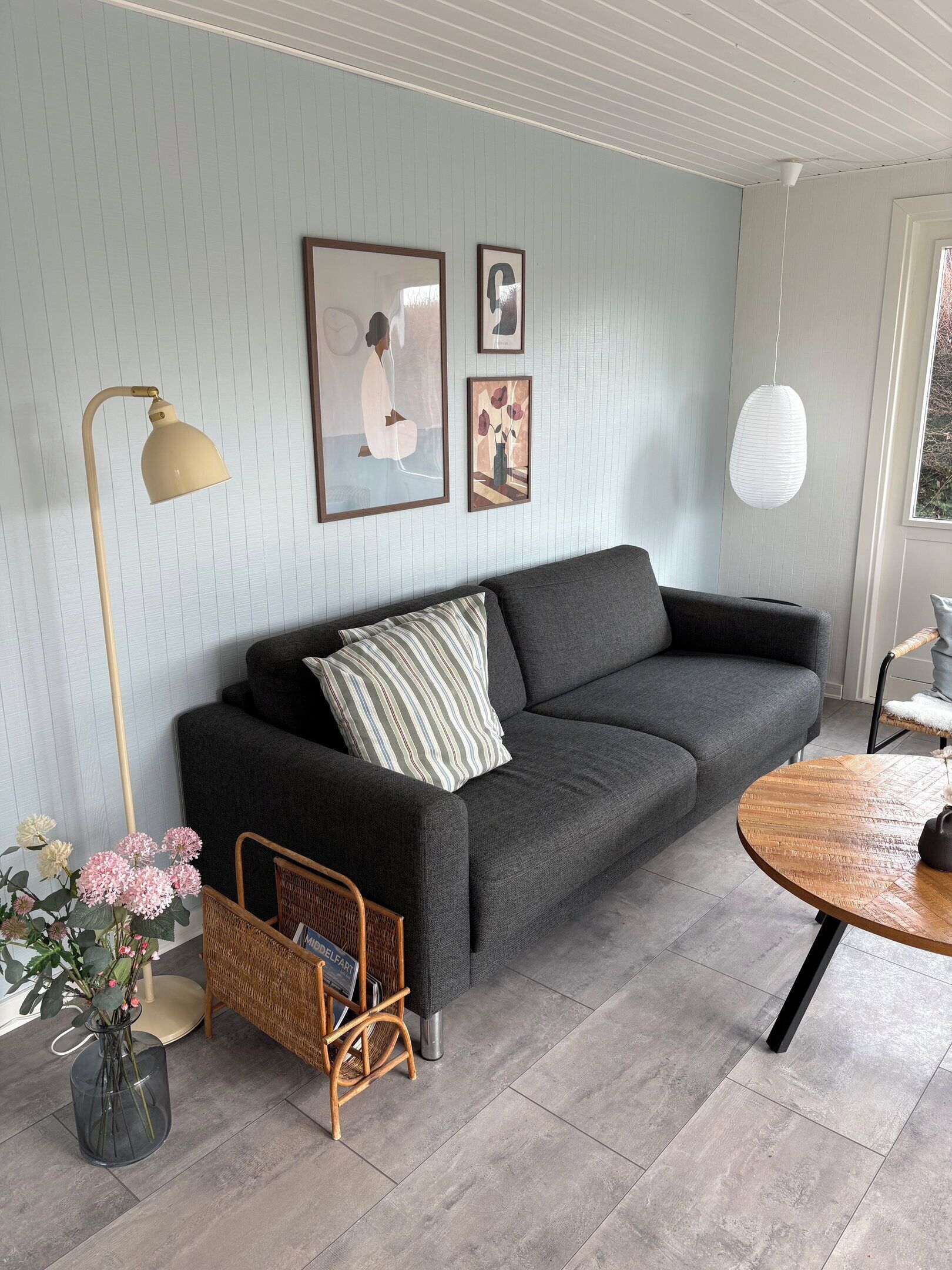 Una sala de estar moderna con un sofá gris, una mesa de centro redonda de madera, una lámpara de pie y cuadros enmarcados en la pared.