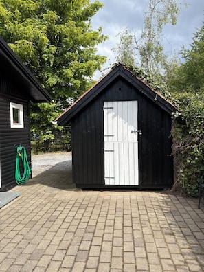 A small black shed with a white door, a green garden hose, and a paved driveway. - Summer house near the sea and good experiences (Børkop)