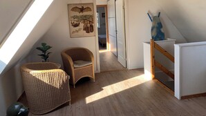 Una habitación con techo inclinado, una silla de mimbre, una pequeña planta en maceta, una ilustración de murciélago en la pared y una escalera que conduce a otra habitación.