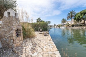 Un sentiero in pietra conduce a uno specchio d'acqua, con una costruzione in pietra a sinistra e palme sullo sfondo.