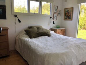 A bedroom with a bed, two bedside lamps, a nightstand with a green bowl, a wooden dresser, and a window with a view of greenery. - Nice summer house with sea view (Jægerspris)