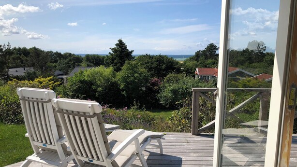 A wooden deck with two white chairs and a glass door leading to a view of a garden and distant buildings. - Nice summer house with sea view (Jægerspris)
