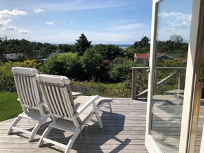 A wooden deck with two white chairs and a glass door leading to a view of a garden and distant buildings. - Nice summer house with sea view (Jægerspris)