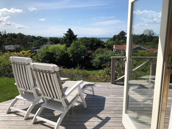 A wooden deck with two white chairs and a glass door leading to a view of a garden and distant buildings.