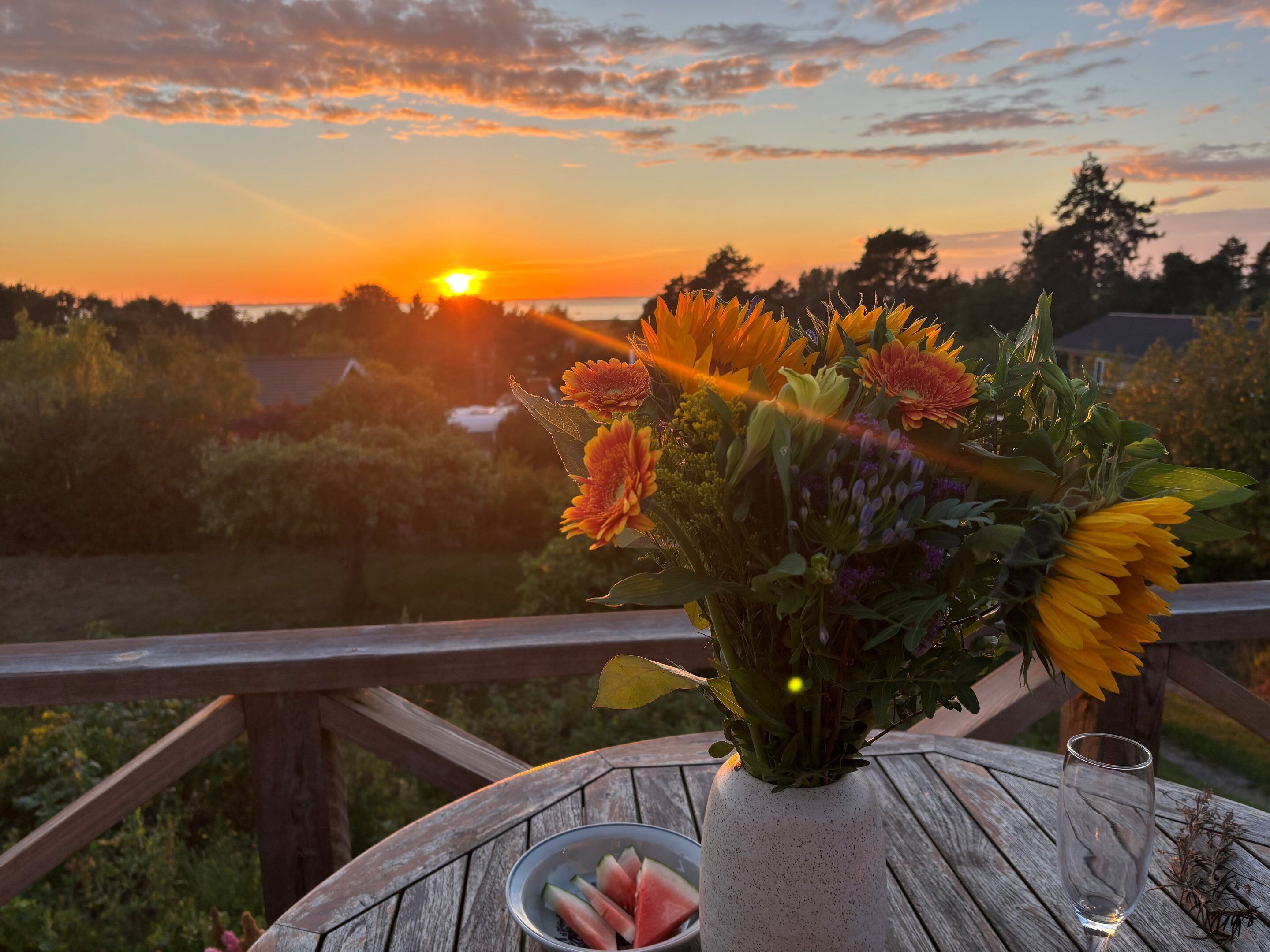 Une vue au coucher du soleil, avec une table sur laquelle on aperçoit un vase de fleurs, un verre et un bol rempli de tranches de pastèque.