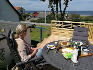 Eine Person sitzt an einem runden Tisch mit Blick auf das Meer, eine Holzterrasse und ein rotes Gebäude im Hintergrund.