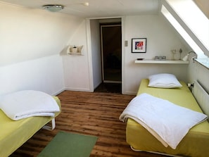 A small, well-lit room with two single beds, a wooden floor, a green mat, a small shelf with toiletries, and a framed picture on the wall.