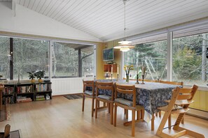 A dining area with a table set for four, surrounded by wooden chairs, a bookshelf, and a television. - Nice summer house in the middle of the forest (Store Fuglede)