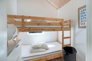 Ein Etagenbett mit Holzbettgestell, einer weißen Matratze und einem Kopfkissen. Darüber befindet sich ein Fenster, an der Wand eine gerahmte Alphabettafel und ein schwarzer Mülleimer.