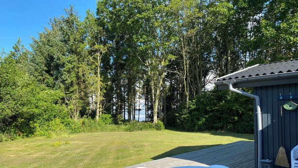 A wooden deck with a table and chairs, surrounded by a forested area and a small shed with a birdhouse. - Idyllic summer house at the water's edge (Spøttrup)