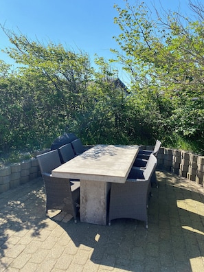 Un patio exterior con una mesa rectangular y sillas de mimbre, rodeado por un muro de piedra y follaje.