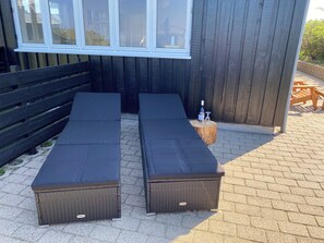 Deux fauteuils de jardin en osier avec des coussins noirs, une table en bois avec une bouteille et des verres, et une clôture en bois.