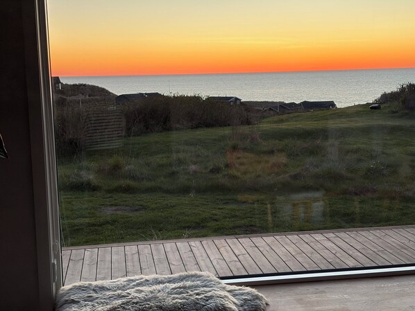 Blick aus einem Zimmer mit großem Fenster auf eine Wiese und ein Gewässer bei Sonnenuntergang.