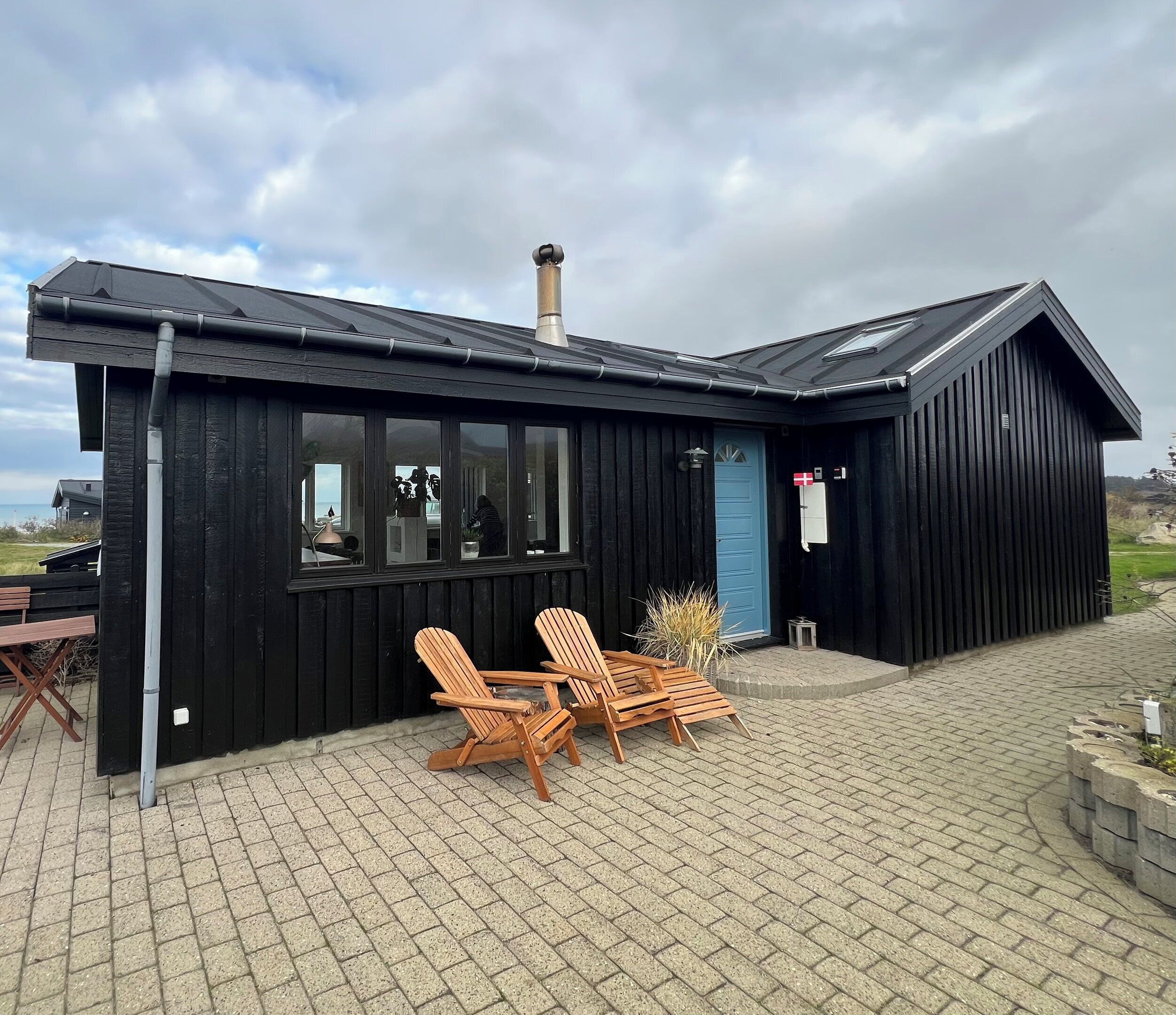 A black wooden building with a chimney, large windows, and a blue door. There are two wooden chairs and a table outside.