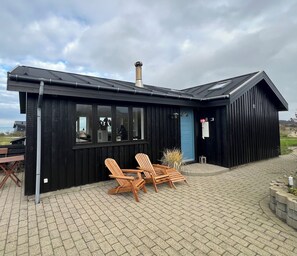 Un bâtiment en bois noir, doté d’une cheminée, de grandes fenêtres et d’une porte bleue. On y trouve deux chaises en bois et une table à l’extérieur.