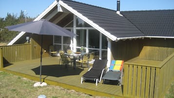 Et hus med en træterrasse, en overdækket terrasse med et bord og stole, samt en stor parasol.