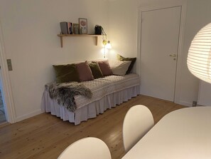 Ein Zimmer mit einem Bett, einem Nachttisch, einer Lampe, einem Regal mit Büchern und einem Bild sowie einer Tür.
