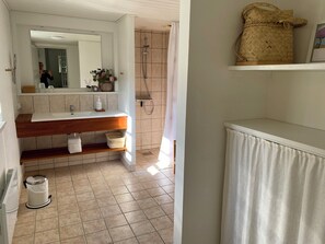 Ein Badezimmer mit einem weißen Waschbecken, Holzregalen, einer Dusche und einem hängenden gewebten Korb.
