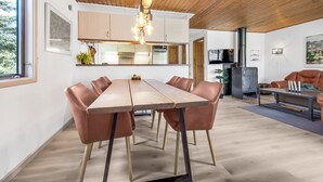 Un intérieur moderne avec un plafond en bois, un coin repas avec une longue table et des chaises en cuir, une cuisine avec des meubles blancs et une cheminée.