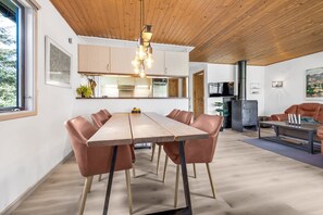 A modern interior with a wooden ceiling, a dining area with a long table and leather chairs, a kitchen with white cabinets, and a fireplace.