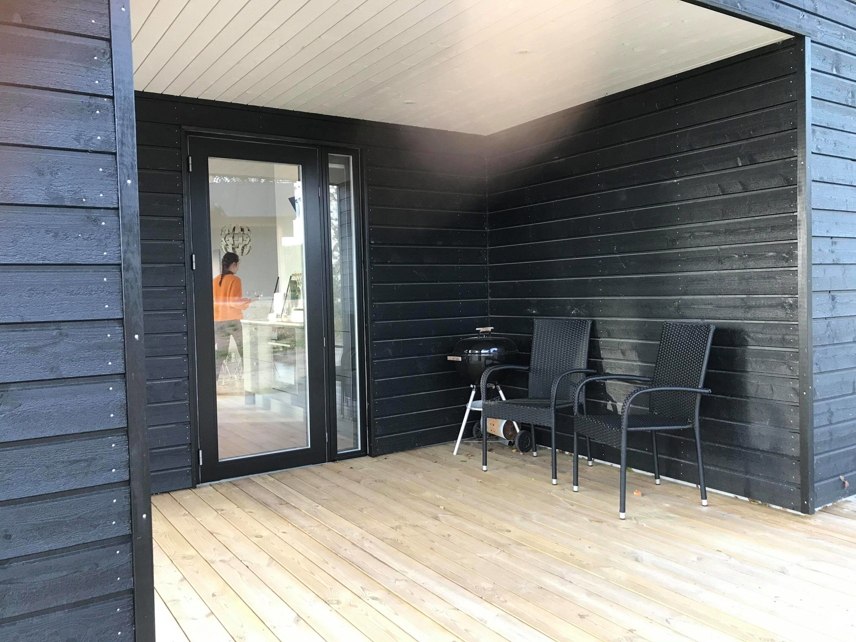 Un terraço de fusta amb dues cadires i una taula, una porta corredissa de vidre i una persona al fons.