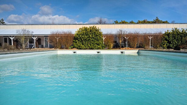 A swimming pool with clear water, surrounded by a building with a covered walkway and trees.