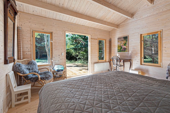 A wooden cabin interior with a large bed, a dining area, and a view of the forest through large windows.