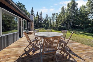 Une terrasse en bois avec une table ronde et des chaises, entourĂ©e de portes vitrĂ©es et offrant une vue sur une forĂȘt.