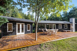 Une terrasse en bois avec des meubles dâextĂ©rieur, un arbre et un bĂątiment dotĂ© de grandes fenĂȘtres et dâune porte.
