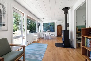 A spacious living area with a large window, a fireplace, a dining table with chairs, and a bookshelf. - Beautiful summer house near forest and beach (Højby)