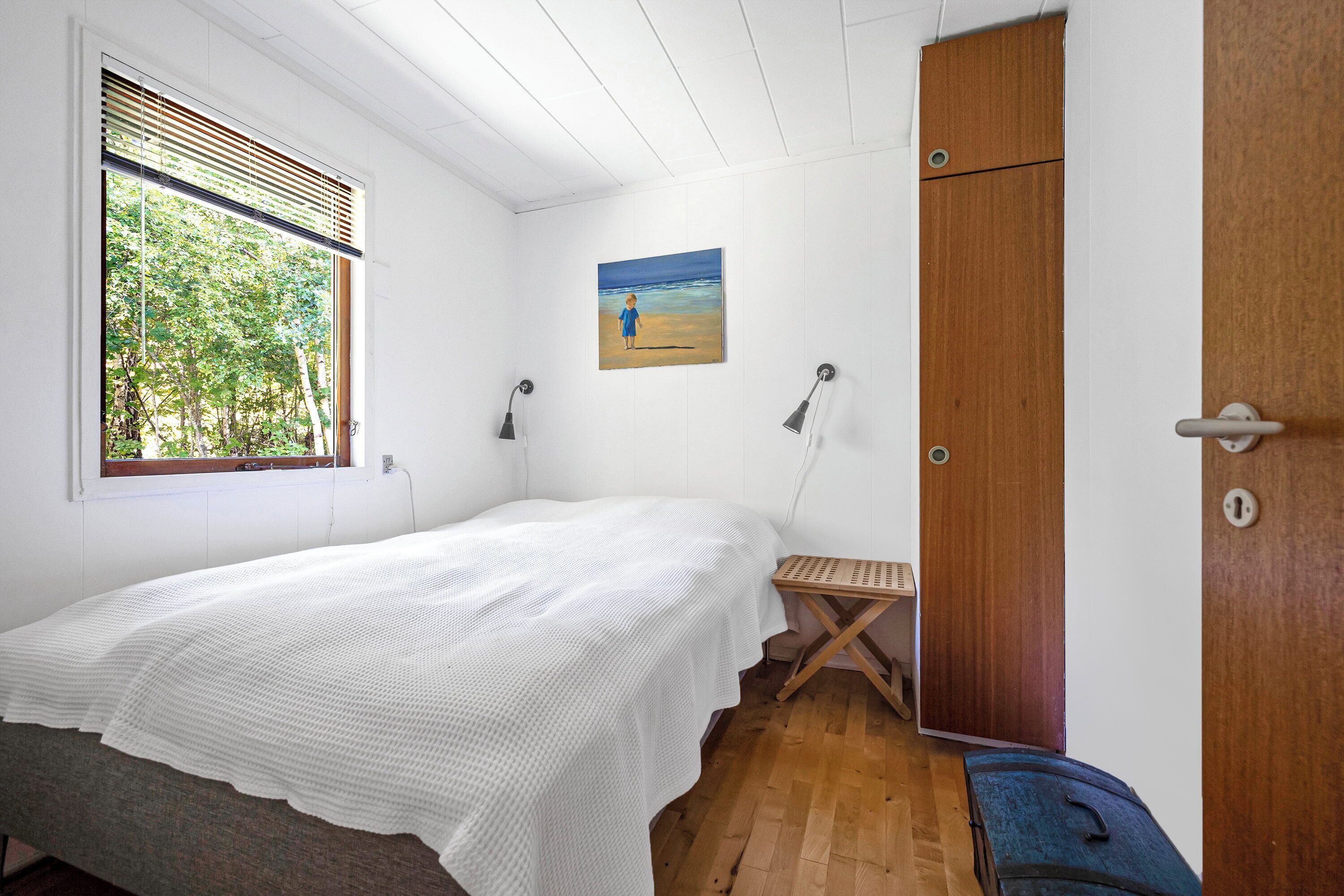 A bedroom with a bed, a wooden door, a window with blinds, a painting on the wall, and a small table.