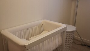 Ein weißes Babybett mit Matratze, ein weißer Wäschekorb und eine Stehlampe in einem Raum mit schlichter Wand und Holzboden.