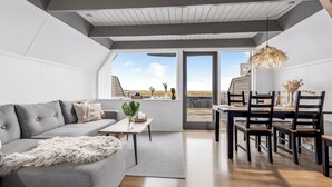 A modern living room with a grey sofa, a wooden dining table, and a view of the outdoors. - Family holiday with sea view in Gedser Feriepark (Gedser)