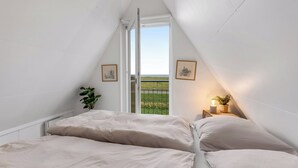 A bedroom with a sloped ceiling, a bed with white bedding, a small bedside table with a lamp, and a window with a view of a green field. - Family holiday with sea view in Gedser Feriepark (Gedser)