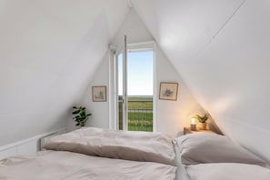 Et soveværelse med skrå væg, en seng med hvide sengetæger, et lille natbord med en lampe og et vindue med udsigt over et grønt marklandskab.