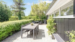 An outdoor patio with a wooden table and chairs, surrounded by greenery and a wooden fence. - Unique holiday home in quiet surroundings (Storvorde)