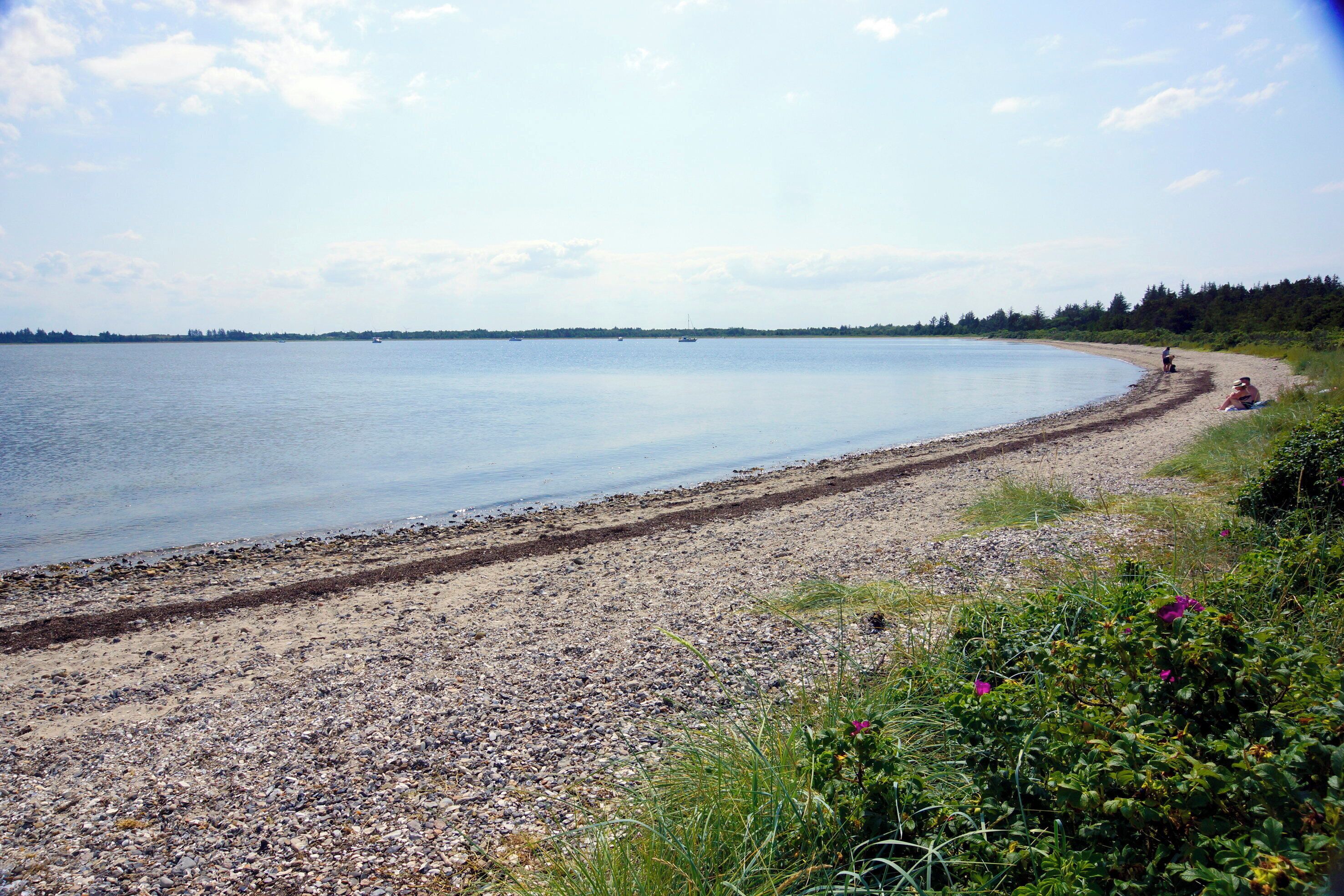 A pebbly beach with a calm sea, a distant shoreline, and a person sitting on the sand.