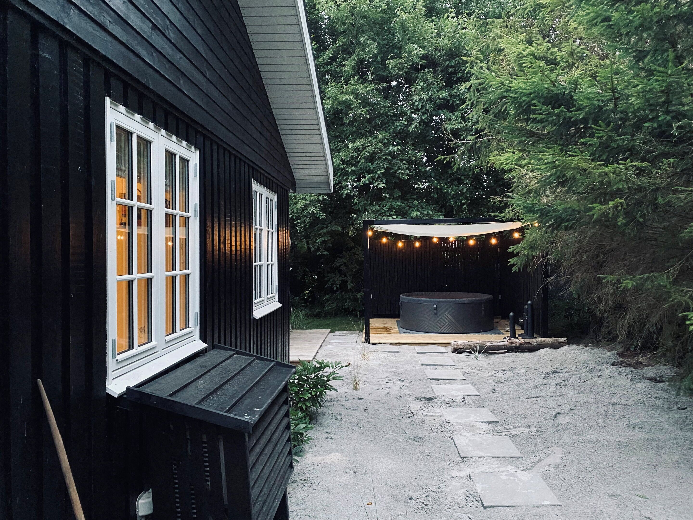 Un bâtiment noir avec des fenêtres blanches, un jacuzzi et des guirlandes lumineuses dans un jardin.