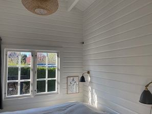 Una habitación con paredes blancas empapeladas, una cama, un ventanal con vistas a zonas verdes y una estructura azul, además de dos lámparas fijadas a la pared.