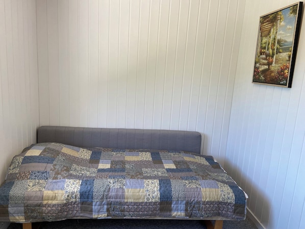 A single bed with a grey headboard and a patchwork quilt, a framed painting on the wall, and a white paneled wall. - Cozy summer house on scenic Djursland (Glesborg)