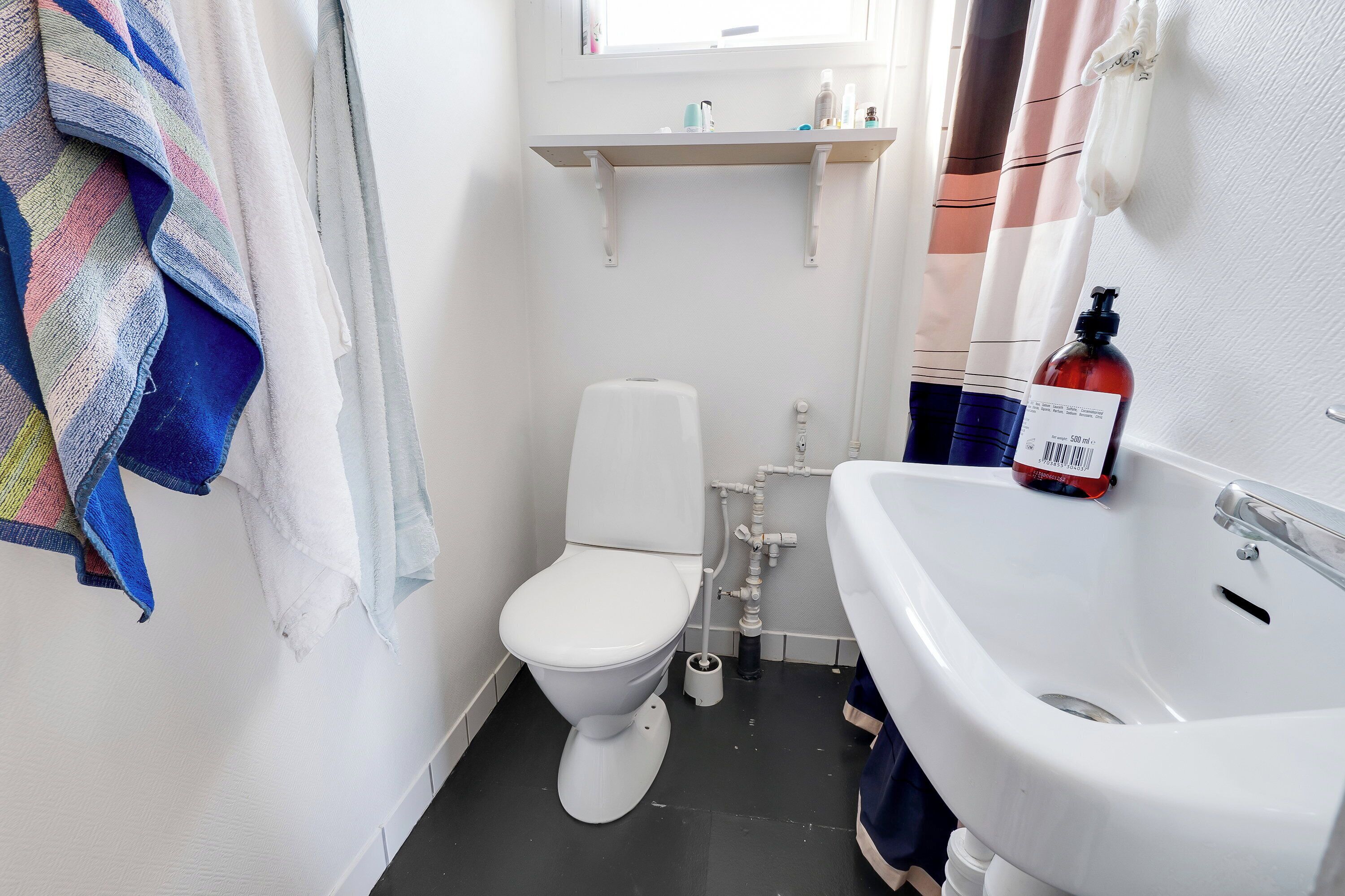 A bathroom with a white toilet, a white sink, and a shower area. There are towels hanging on the wall and a bottle of hand soap on the sink.