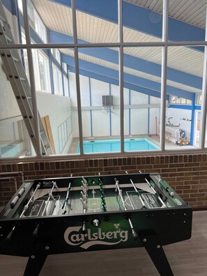 A foosball table with a 'Carlsberg' logo, a brick wall, and a view of a swimming pool through large windows.