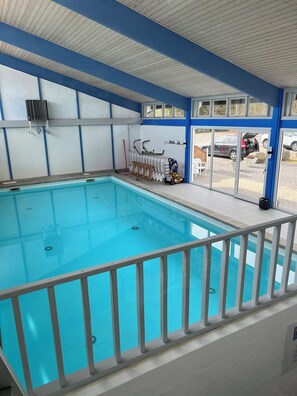 An indoor swimming pool with a white railing, a ceiling with horizontal beams, and a view of parked cars outside. - Cozy holiday apartment close to the beach (Nexø)